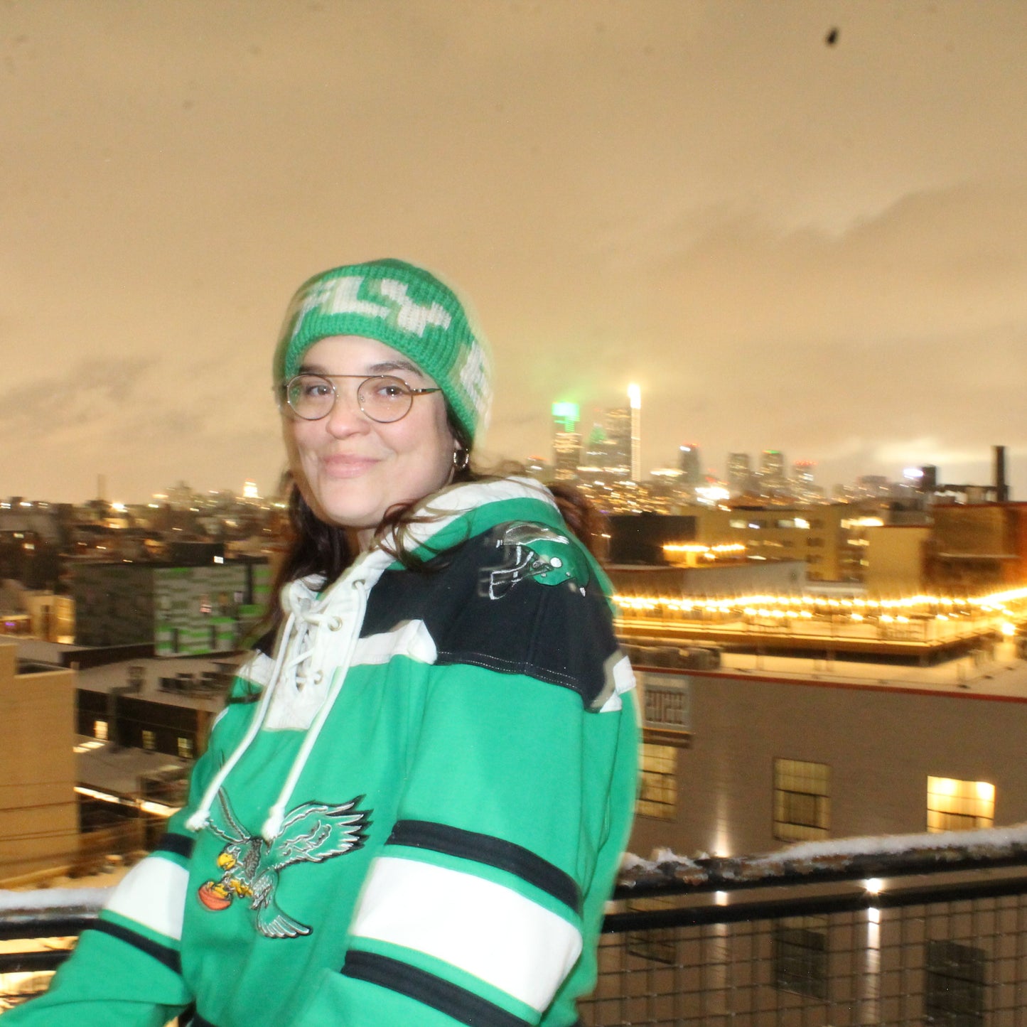 Blurry photo of woman wearing kelly green and white beanie with the word FLY standing in front of the Philadelphia skyline