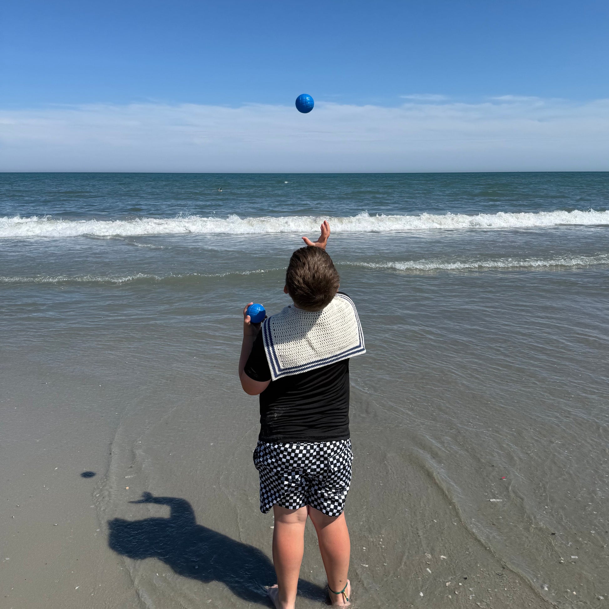 A child standing on a sandy beach facing the ocean and tossing a blue bocce ball in the air. They are wearing a black top, a white crochet sailor scarf draped over their shoulders, and black-and-white patterned shorts under a clear blue sky.