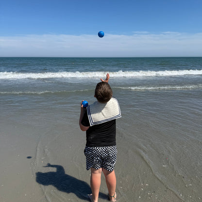 A child standing on a sandy beach facing the ocean and tossing a blue bocce ball in the air. They are wearing a black top, a white crochet sailor scarf draped over their shoulders, and black-and-white patterned shorts under a clear blue sky.