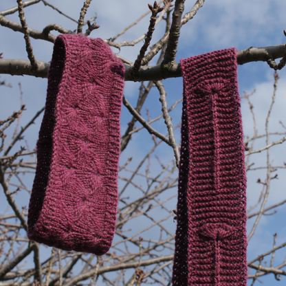 Two purple knitted garments, a headband and a scarf, both with fleabane flower motifs and hanging on a tree branch with a blue sky background.