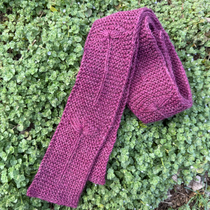 Purple knitted scarf on green grass and clovers, featuring a flower motif