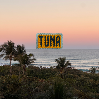 Crochet piece with 'TUNA' on a beach at sunset