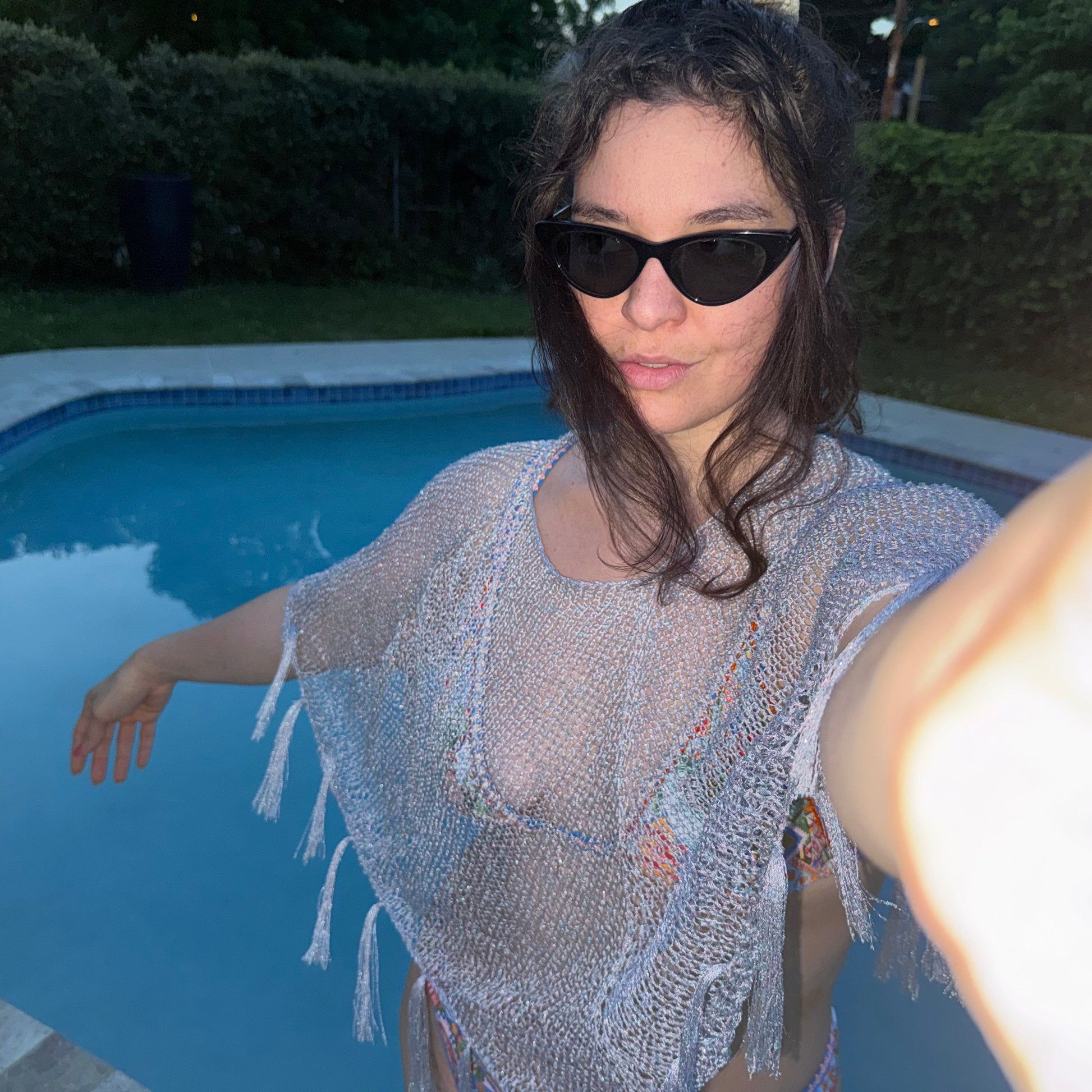 Woman in a silver handknit poncho and sunglasses by a pool at night.