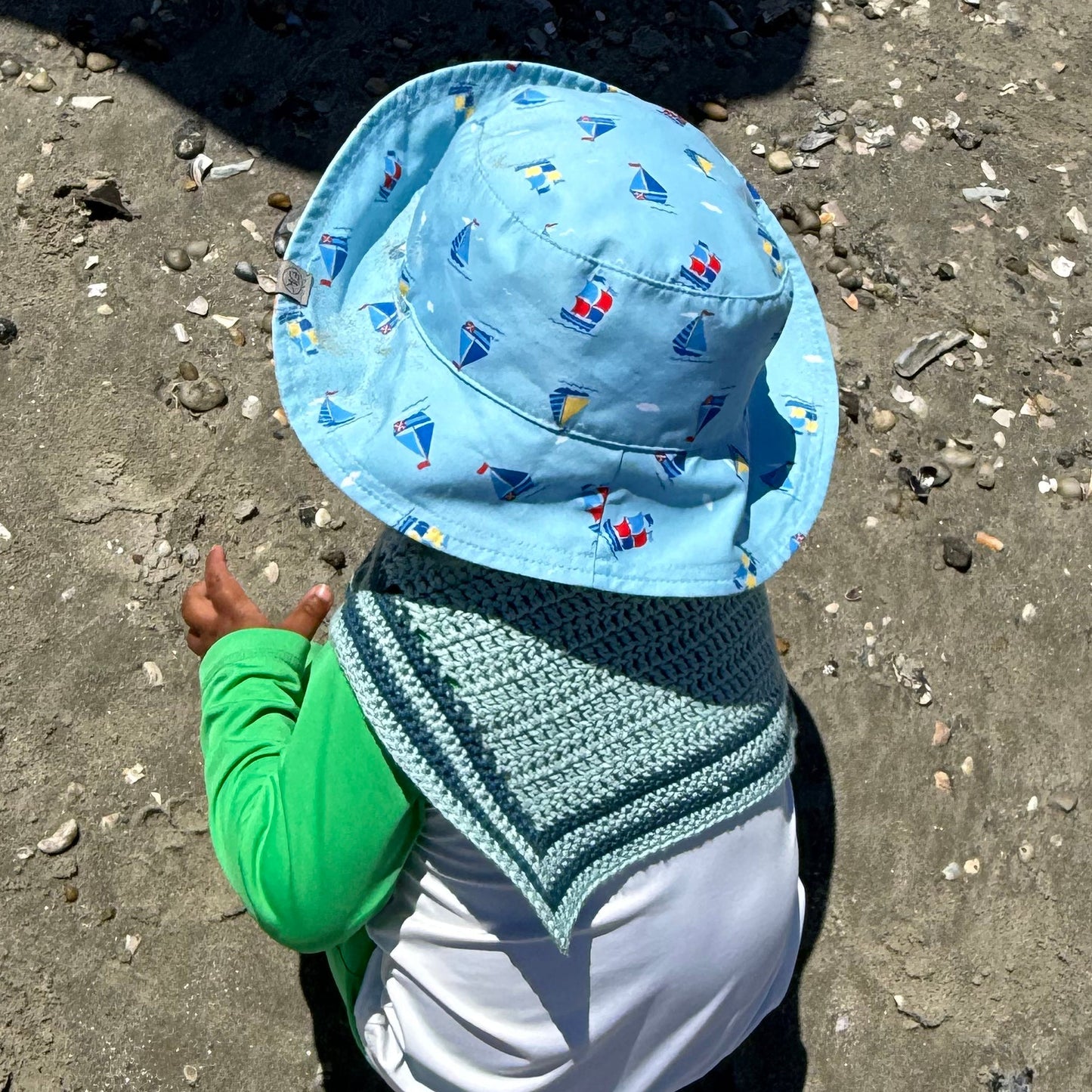 A child wearing a light blue sun hat with a sailboat pattern and a matching crocheted sailor scarf playing on a sandy beach.
