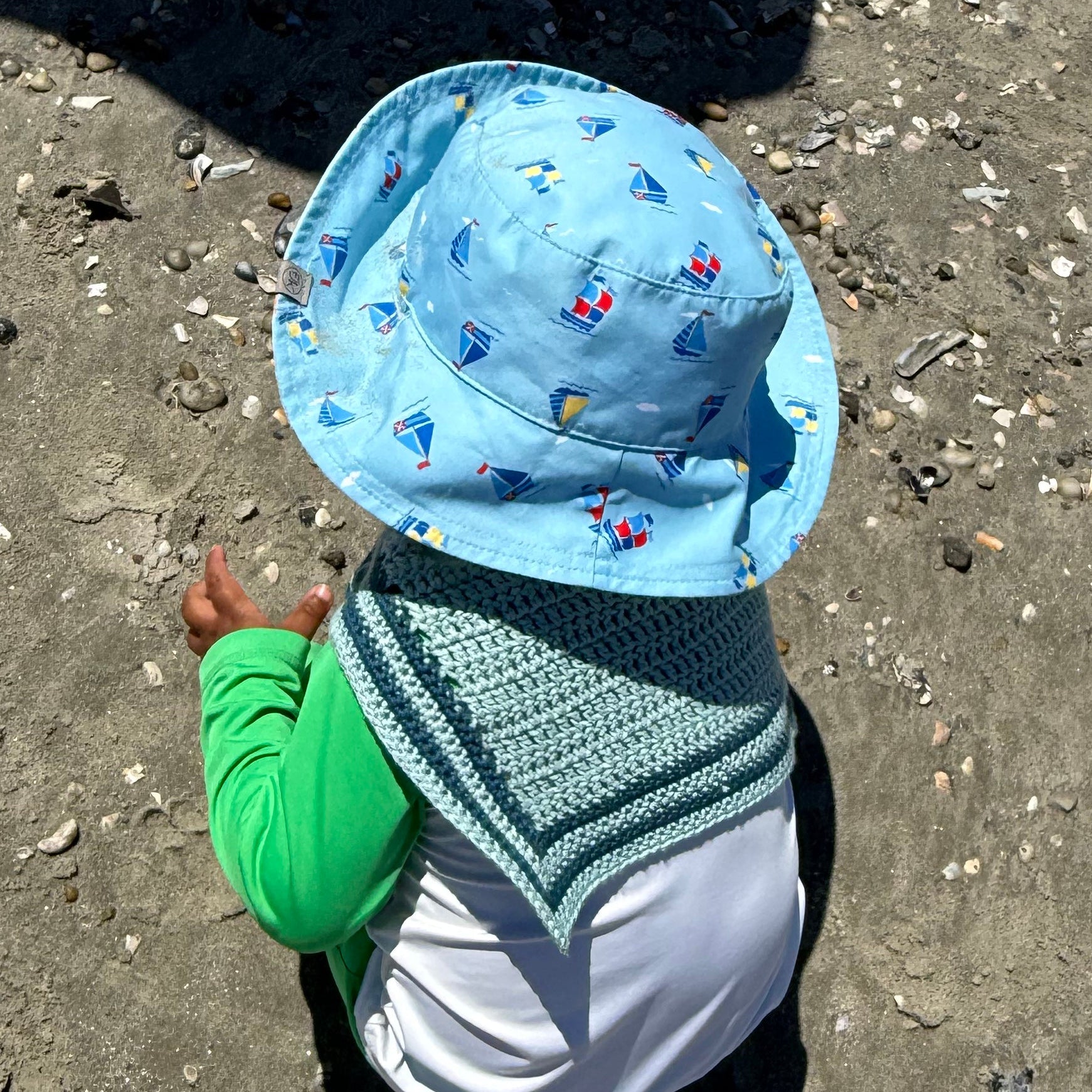 A child wearing a light blue sun hat with a sailboat pattern and a matching crocheted sailor scarf playing on a sandy beach.