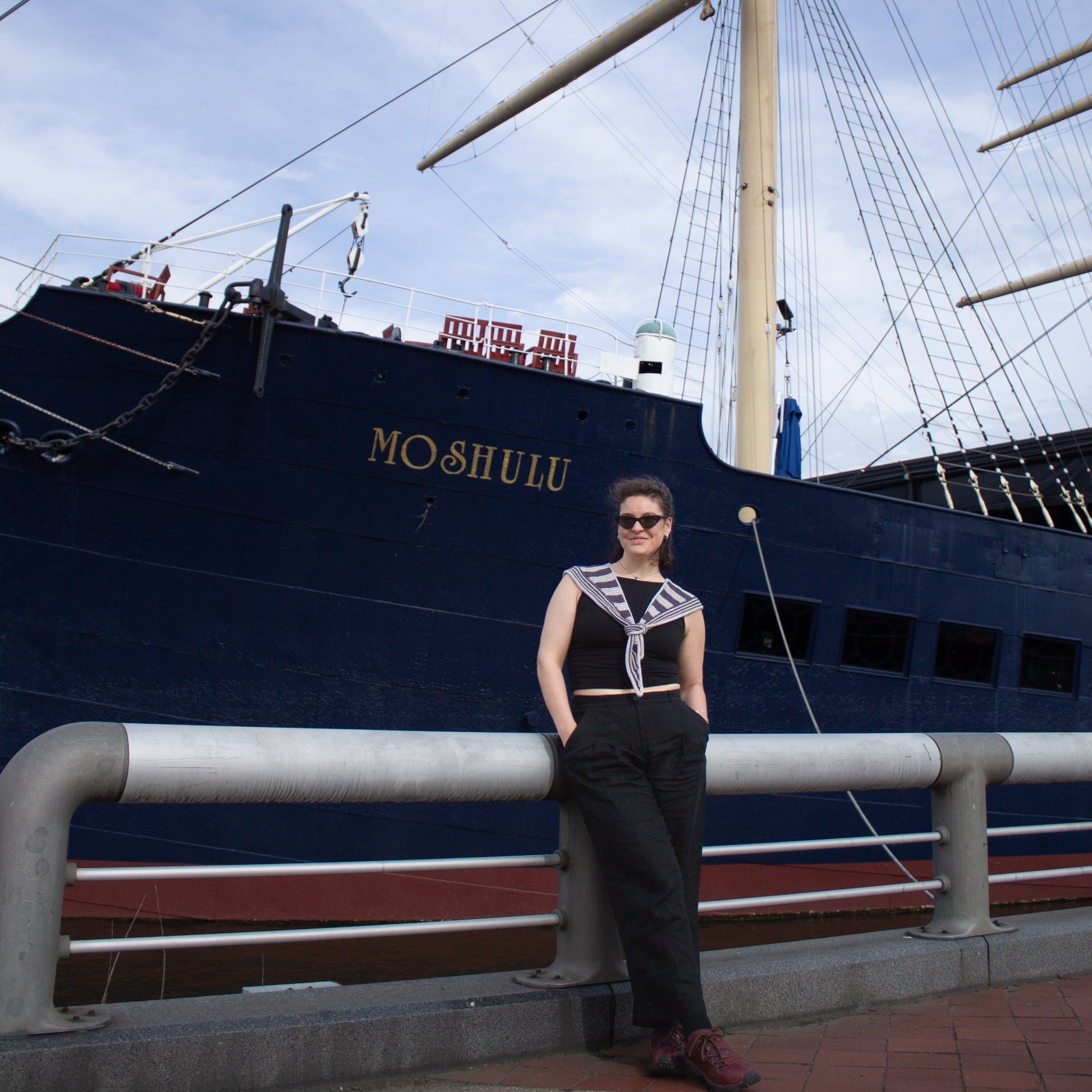 Person wearing a striped hand knit scarf standing in front of a large ship named 'Moshulu' under a blue sky.