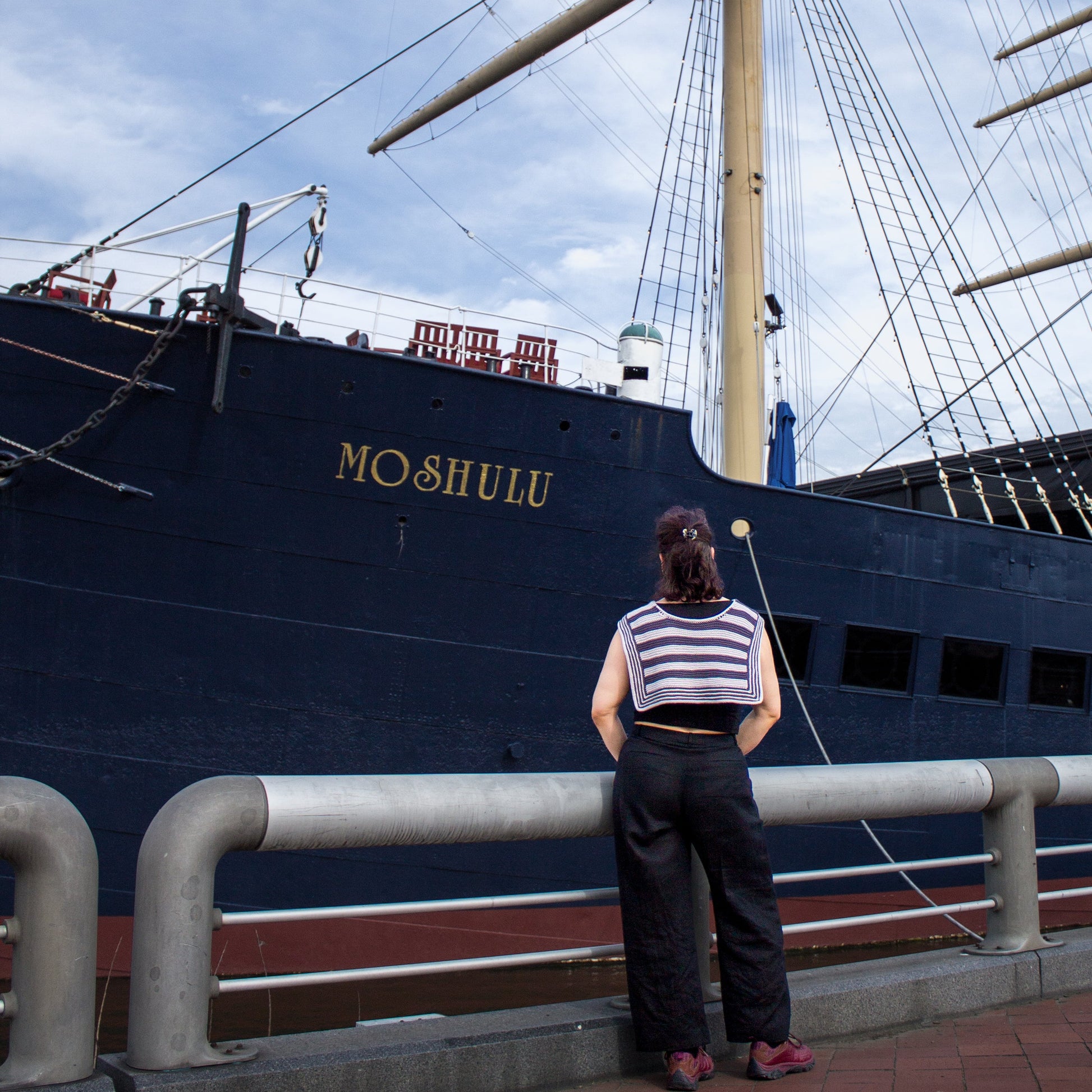 Person in a handknit striped scarf standing in front of a large ship named 'Moshulu' under a blue sky.